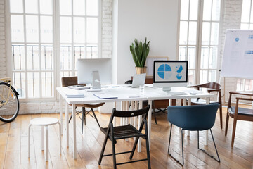 Empty modern office with coworking desk and flip chart for presentation, no people around. Renovated loft interior design workplace with computers and gadgets on table. Freelance workspace concept.