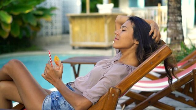 Sexy young woman with drink relaxing on sunbed by pool