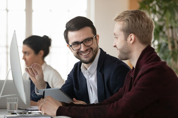 Smiling male colleagues brainstorm work together on computer in coworking space, discuss business ideas in group. Happy men coworkers cooperate using gadgets in office. Collaboration concept.