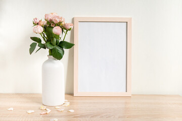 Portrait white picture frame mockup on wooden table. Modern ceramic vase with roses. White wall...
