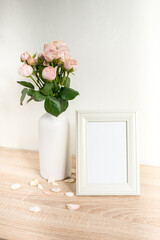 Portrait white picture frame mockup on wooden table. Modern ceramic vase with roses. White wall background. Scandinavian interior. 
