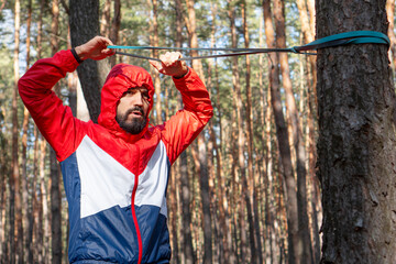 Athletic bearded man pulls the resistance band in a morning forest. Exercises on a fresh air during...