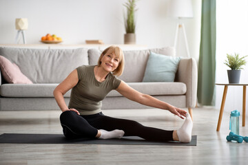Domestic workout. Cheerful senior lady stretching her leg on sports mat in living room, empty space