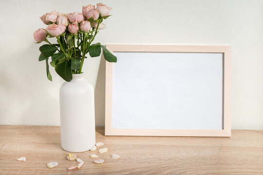 Portrait white picture frame mockup on wooden table. Modern ceramic vase with roses. White wall background. Scandinavian interior. 
