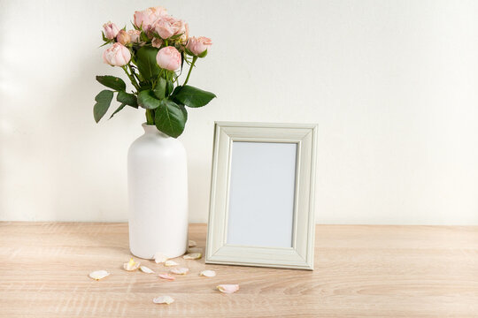 Portrait white picture frame mockup on wooden table. Modern ceramic vase with roses. White wall background. Scandinavian interior. 

