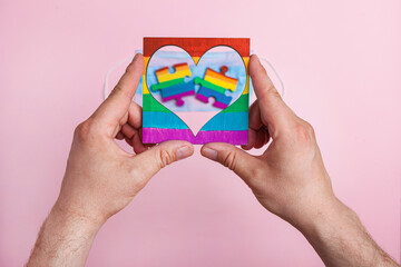 LGBT symbol frame heart in male hands inside puzzles painted by a rainbow on a medical mask. Coronavirus quarantine, online festival and International Day Against Homophobia.