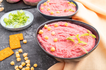 Hummus with beet and microgreen basil sprouts in ceramic bowl on a black concrete background. Side view, close up.