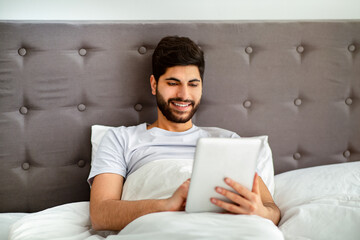 Weekend leisure concept. Relaxed arab man using digital tablet, sitting in bed and surfing internet online