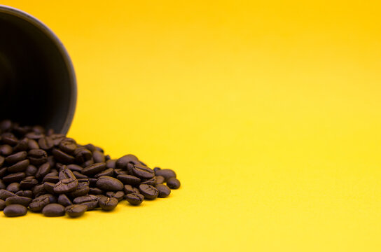 Coffee  beans coming out of an upturned cup on a table and yellow wall in the background. Horizontal image in the left third, close-up shot. Vivid an calid colors.