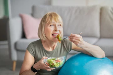 Poster Sports and nutrition concept. Smiling senior lady leaning on fitness ball, eating fresh vegetable salad at home © Prostock-studio
