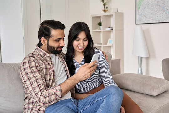 Happy Young Indian Family Couple Using Smart Phone Looking At Cellphone Sit On Sofa Together Relaxing At Home. Smiling Husband And Wife Checking Social Media Apps Or Shopping In Mobile Application.