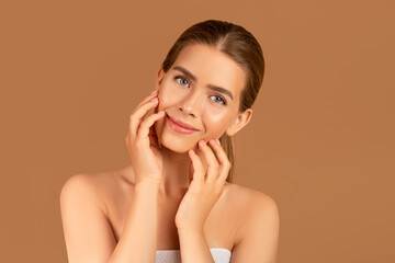 Sexy young woman touching perfect smooth skin on her face, looking at camera on brown background. Wellness and spa