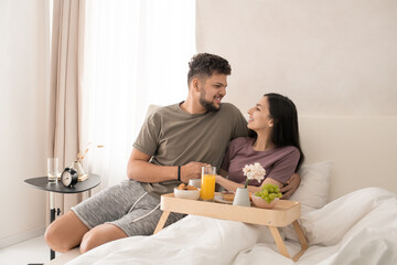 Obraz na płótnie Canvas Happy young affectionate couple looking at one another with smiles while sitting in bed, having tasty breakfast and discussing something nice