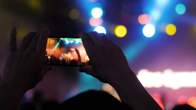 Close up: man hands silhouette taking photo or recording video of live music concert with smartphone at nightclub. Bright colorful stage lighting. Photography, technology concept