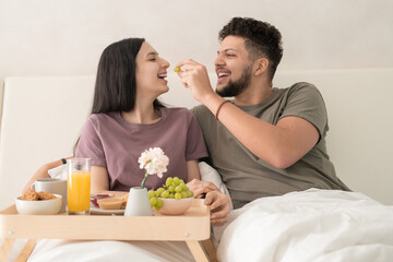 Obraz na płótnie Canvas Young cheerful man putting grape in his wife mouth while both sitting under white blanket in double bed and enjoying breakfast