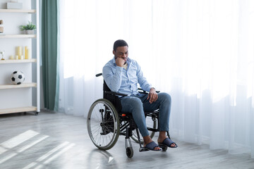 Full length of impaired black man sitting in wheelchair near window, feeling sad and desperate at...
