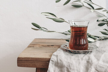 Close-up of Turkish black tea in glass on silver saucer. Ramadan Kareem greeting card, invitation. Green olive tree branches on old wooden table background. Muslim Iftar dinner concept. Front view.