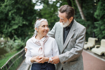 Happy senior man and charming lady smiling and hugging outdoors. Lovely mature family dressed in stylish clothes walking together at green park.