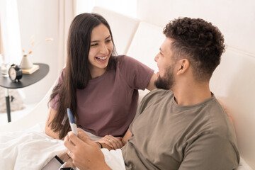 Fototapeta premium Young laughing brunette female embracing her husband holding positive pregnancy test while both sitting in bed and looking at one another