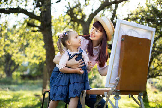 Mother's Day, child art and creativity concept. Outdoor portrait of pretty young mom and cute 3-aged little daughter, painting on easel a picture in a beautiful green park or garden at spring.