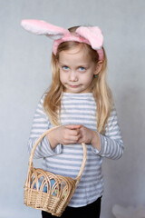 blonde girl in a striped jacket, on a bright background, with rabbit ears on her head, holding a basket of eggs, smiling angrily looks into the frame