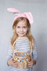 blonde girl in a striped jacket, on a bright background, with rabbit ears on her head, holding a basket of eggs, smiling looks into the frame