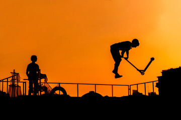 Unrecognizable teenage boy silhouette showing high jump tricks on scooter against orange sunset sky...