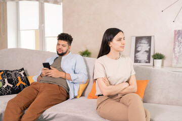 Young dissatisfied woman in casualwear crossing arms by chest while sitting on couch against her husband scrolling in smartphone
