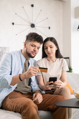 Young bearded man holding credit card while his wife with smartphone entering its number while going to pay for ordered goods or service