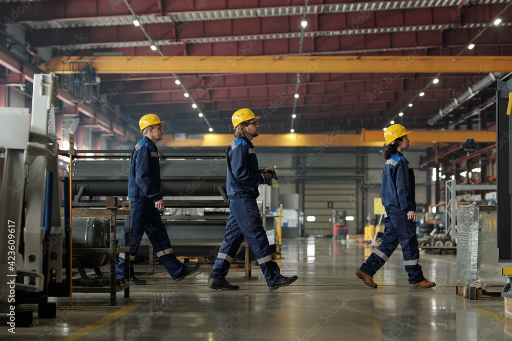 Poster side view of three engineers of modern factory or industrial plant moving forwards while going to an