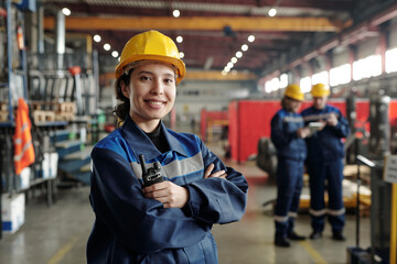 Young smiling female worker of modern industrial plant or factory in workwear and protective helmet standing in large workshop