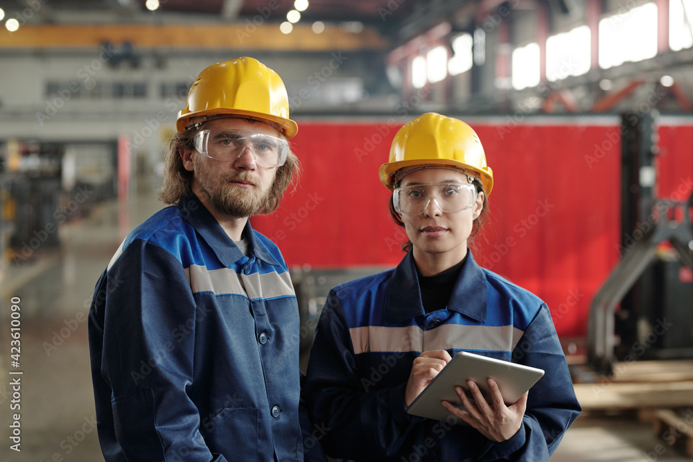 Poster two young serious engineers in protective hardhats and workwear looking at you while standing in fro
