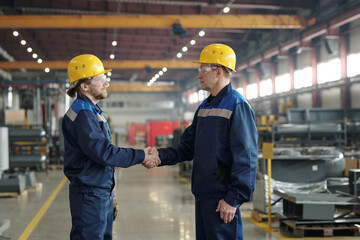 Two male engineers in protective hardhats and workwear greeting one another by handshake inside...