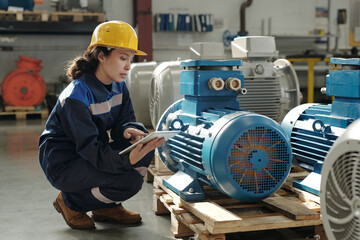 Fototapeta na wymiar Young serious female engineer in protective helmet and workwear squatting by new industrial equipment and using digital tablet