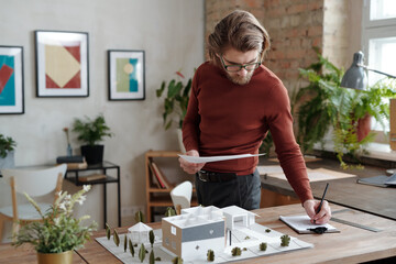 Young serious architect in casualwear and eyeglasses making notes while standing by table with model of new house and yard in office