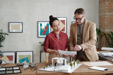 Young female architect and her mature male colleague holding paper over model of new house and yard while discussing details of sketch