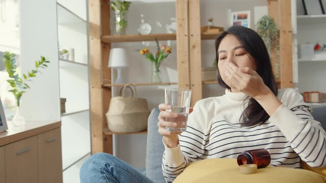 Sick young Asia woman holding pill glass of water take medicine sit on couch at home. Girl taking medicine after doctor order, quarantine at house, Coronavirus social distancing quarantine concept.