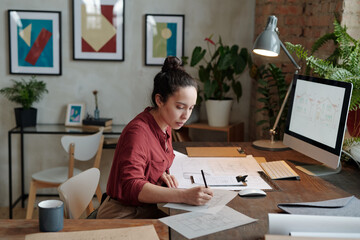 Young serious female architect with pencil drawing sketch on paper while sitting by table in front of computer screen with picture of house