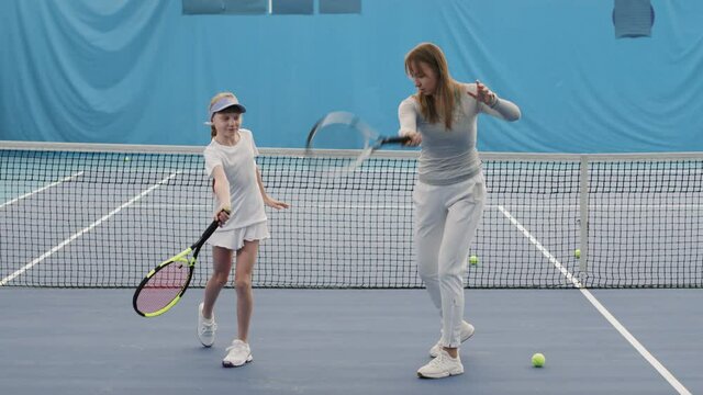 Slow-motion full shot of young female tennis coach teaching little girl how to throw ball with tennis racket training together on court indoors