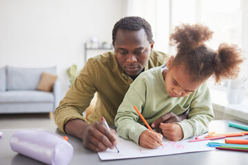 Portrait of caring African-American father and daughter drawing together while sitting at desk in home interior