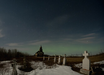 Bleak looking dilapidated graveyard with an old church in the background under a starry night.  Scattered clouds and weak Aurora can be seen in the sky.
