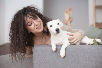 Cheerful curly woman kissing hugging little dog feeling love tenderness at cozy minimalistic home