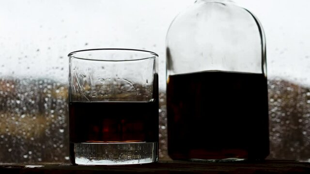 A glass of whiskey with a bottle on the bar near the rain window. Drops run down the glass. Alcohol at the bar.