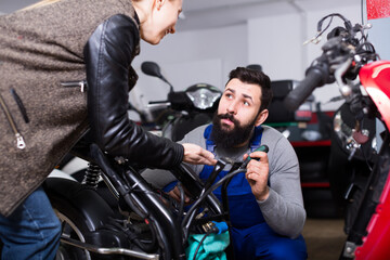 Fototapeta na wymiar friendly male worker assisting blonde female customer in repairing motorcycle in workshop
