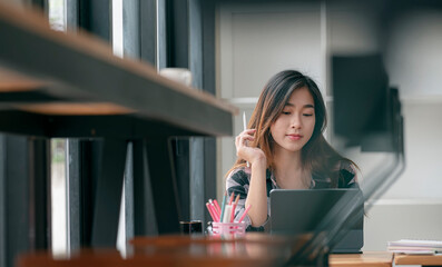 Portrait of young asian woman holding stylus pen while working on tablet computer at modern cafe.