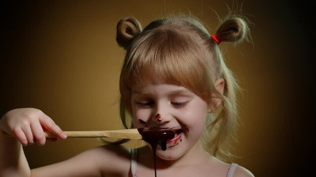 Satisfied face of pretty Caucasian child enjoying unhealthy sweet food indoors. Happy little girl licking melted chocolate isolated on dark background. Joyful kid eating dessert sweet candy syrup