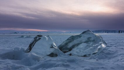 Large transparent shining ice hummocks on a frozen and snowy lake. Close-up. People walking on the ice can be seen in the distance. Sunset pink sky. The outlines of the mountains. Baikal