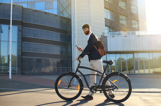Young businessman in protective mask with bicycle and smartphone on city street.