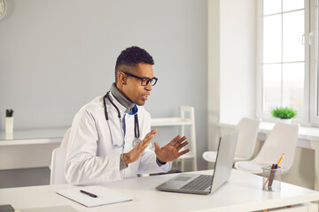 Young black man doctor therapist sitting, talking online with patient and explaining something with hands during videocall from clinic office. Telemedicine and online medicare concept