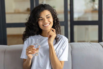 Portrait happy young african american woman smiling sit on couch holding glass of still or mineral...